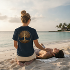 A woman on a sandy beach kneels beside a man lying down, wearing a navy blue t-shirt with a large gold tree-of-life graphic on the back. The background features a calm ocean, tropical palm trees, and a soft sunset sky.
